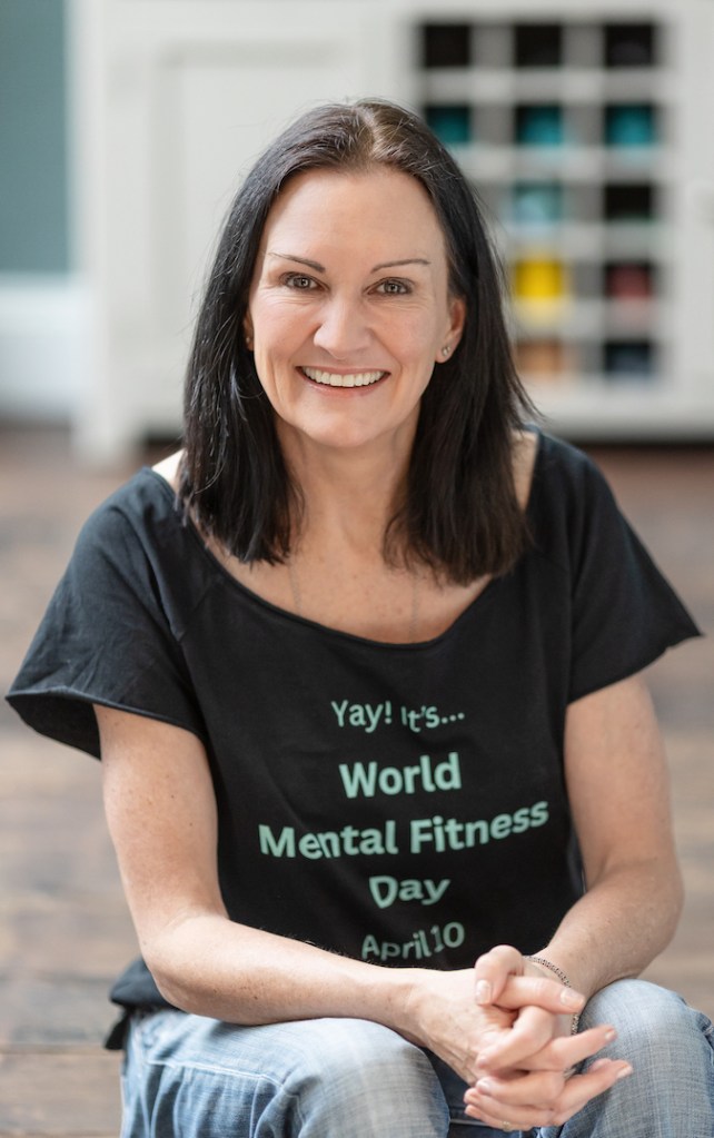 A smiling woman sitting on the floor, wearing a black shirt that says 'Yay! It's... World Mental Fitness Day April 10'.
